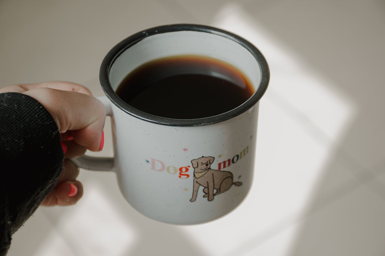 Home A hand holds a Dog Mom mug filled with coffee, with sunlight casting shadows.